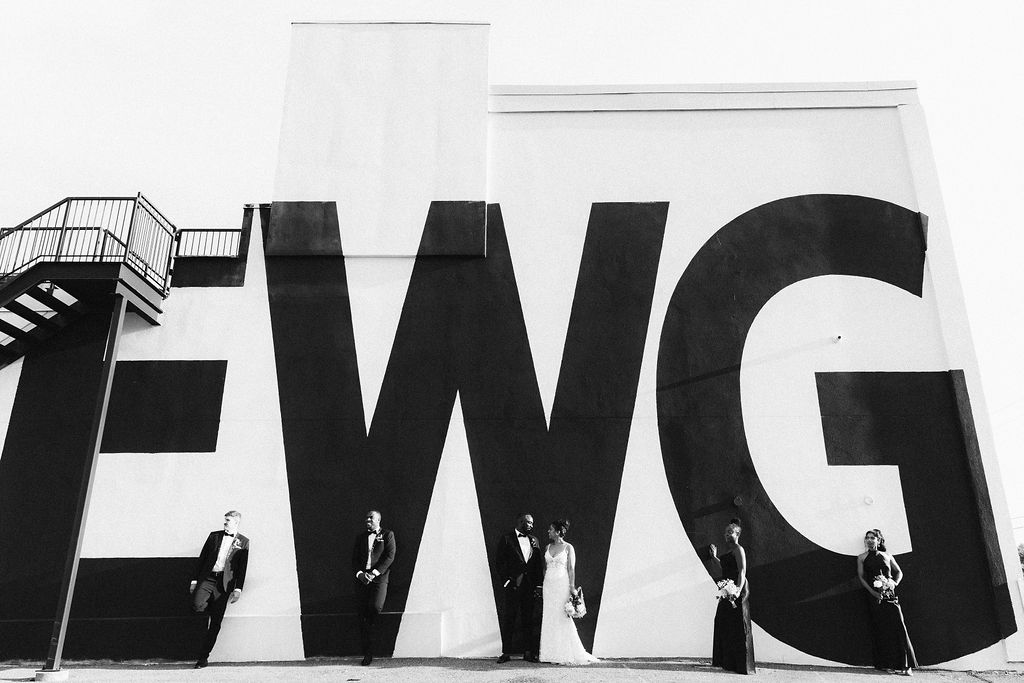 Wedding party posing with EWG graphic on the exterior wall at Eglinton West Gallery wedding, black and white